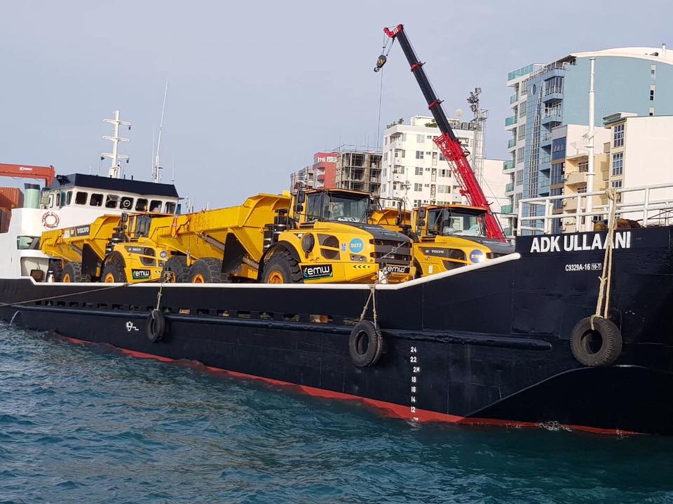 Landing craft alongside at Malé carrying three articulated dump trucks, with a mobile crane on the quay